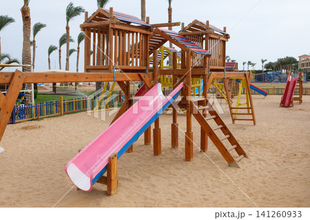 Wooden playground equipment features a pink slide and climbing elements in a sandy area, with palm trees and colorful fencing in the background Wooden playground equipment features a pink slide and climbing elements in a sandy area, with palm trees and colorful fencing in the background 141260933