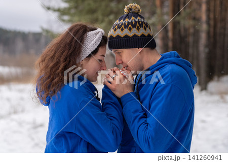 Outdoor happy couple in love posing in cold winter weather. A man and a woman in blue hoodies. Emotional young couple having fun while walking by winter forest, loving man hugging his laughing woman Outdoor happy couple in love posing in cold winter weather. A man and a woman in blue hoodies. Emotional young couple having fun while walking by winter forest, loving man hugging his laughing woman 141260941