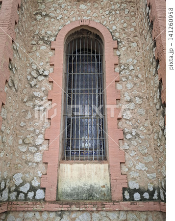 A tall, arched stained glass window sits in a stone wall of an abandoned church in Spain. The window is behind a rusted iron security grate. The church shows signs of age and neglect. A tall, arched stained glass window sits in a stone wall of an abandoned church in Spain. The window is behind a rusted iron security grate. The church shows signs of age and neglect. 141260958