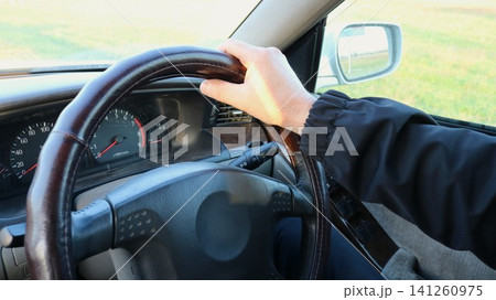 view from inside the car: a driver in a dark windbreaker holds the top of the steering wheel. The speedometer, tachometer, and air vent are visible, and the roadside is reflected in the outside mirror 141260975