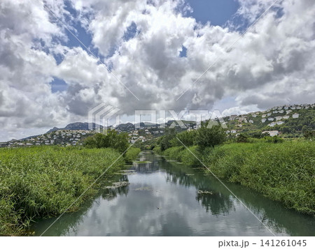 A waterway flows through the Pego wetland region, surrounded by green vegetation and hills. White houses are visible in the distance under a cloudy sky. A waterway flows through the Pego wetland region, surrounded by green vegetation and hills. White houses are visible in the distance under a cloudy sky. 141261045