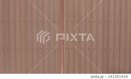 A brown fence made of vertical planks with a textured wood grain. The planks are screwed to the metal frame with even rows of screws, and a vertical joint is visible in the center of the frame. A brown fence made of vertical planks with a textured wood grain. The planks are screwed to the metal frame with even rows of screws, and a vertical joint is visible in the center of the frame. 141261428