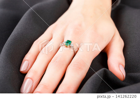 Beautiful female hand with gold ring with diamonds and emerald on gray background Beautiful female hand with gold ring with diamonds and emerald on gray background 141262220