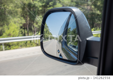 A side mirror captures the view of a winding road lined with green trees under a clear blue sky during a sunny day, highlighting the journey ahead. 141262558