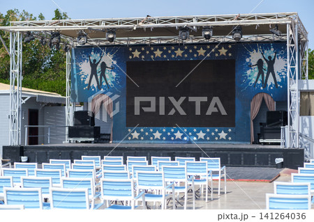 Outdoor stage features a large screen and decorative backdrop, with rows of white chairs arranged for an audience in a sunny setting Outdoor stage features a large screen and decorative backdrop, with rows of white chairs arranged for an audience in a sunny setting 141264036