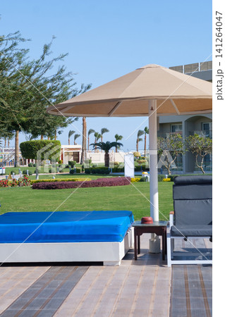 Outdoor lounge area with a blue sunbed and a black reclining chair beneath a large umbrella, surrounded by a well-maintained garden Outdoor lounge area with a blue sunbed and a black reclining chair beneath a large umbrella, surrounded by a well-maintained garden 141264047