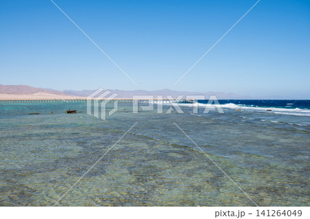 Clear blue waters in a coastal region with a wooden pier extending into the sea, surrounded by mountains under a bright blue sky 141264049