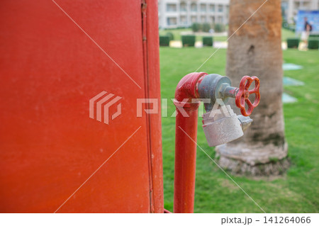 Red fire hydrant valve features a metal handle and protective cap, situated on a green lawn adjacent to a building in an outdoor environment Red fire hydrant valve features a metal handle and protective cap, situated on a green lawn adjacent to a building in an outdoor environment 141264066