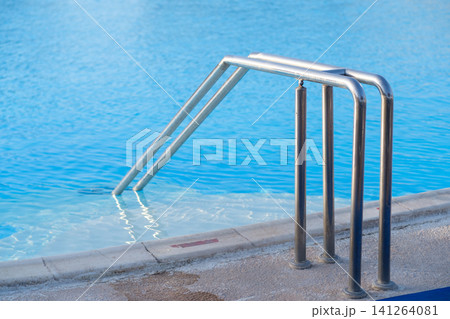 Stainless steel ladder descends into a clear blue swimming pool, featuring a tiled edge and reflections of sunlight on the water surface Stainless steel ladder descends into a clear blue swimming pool, featuring a tiled edge and reflections of sunlight on the water surface 141264081