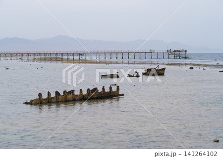 Shipwreck remains are visible in shallow water near a wooden pier, with mountains in the background and a cloudy sky above Shipwreck remains are visible in shallow water near a wooden pier, with mountains in the background and a cloudy sky above 141264102