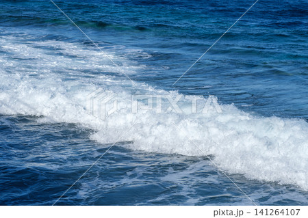 Ocean waves break on the shoreline, producing white foam and ripples, with a vast expanse of deep blue water and clear sky above Ocean waves break on the shoreline, producing white foam and ripples, with a vast expanse of deep blue water and clear sky above 141264107