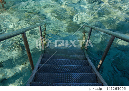 Metal steps descend into clear turquoise water, revealing rocky underwater terrain and sunlight creating patterns on the ocean floor Metal steps descend into clear turquoise water, revealing rocky underwater terrain and sunlight creating patterns on the ocean floor 141265096