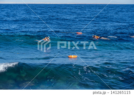 Two individuals are snorkeling in the clear blue ocean, wearing life vests, with buoy markers and a coral reef visible in the background Two individuals are snorkeling in the clear blue ocean, wearing life vests, with buoy markers and a coral reef visible in the background 141265115