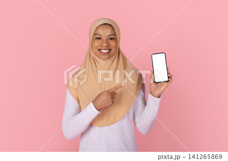 Smiling Black Muslim woman in beige hijab pointing to blank smartphone screen Smiling Black Muslim woman in beige hijab pointing to blank smartphone screen 141265869
