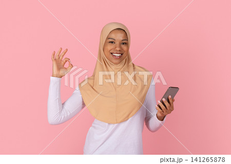 Black Muslim woman in hijab holding smartphone and making approval ok gesture Black Muslim woman in hijab holding smartphone and making approval ok gesture 141265878