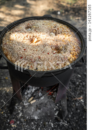 A traditional pot simmering with a flavorful rice dish, filled with spices and ingredients, is placed over an open flame outdoors, surrounded by nature. A traditional pot simmering with a flavorful rice dish, filled with spices and ingredients, is placed over an open flame outdoors, surrounded by nature. 141267165