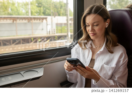 A woman sits comfortably on a train, enjoying music through her earphones while checking her smartphone as the scenery passes by outside the window. A woman sits comfortably on a train, enjoying music through her earphones while checking her smartphone as the scenery passes by outside the window. 141269157