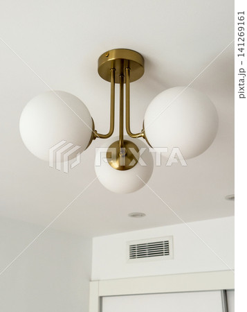 A brass ceiling light with white glass globe shades near a ceiling corner. Modern home lighting in a bright room A brass ceiling light with white glass globe shades near a ceiling corner. Modern home lighting in a bright room 141269161