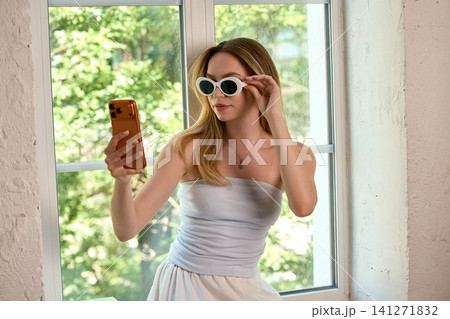 Woman trying white sunglasses while checking appearance with smartphone at home. 141271832
