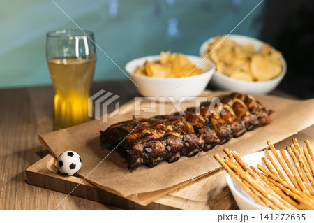 Grilled ribs on a wooden board, beer and snacks arranged for a soccer viewing party, creating a cozy football night atmosphere. Grilled ribs on a wooden board, beer and snacks arranged for a soccer viewing party, creating a cozy football night atmosphere. 141272635