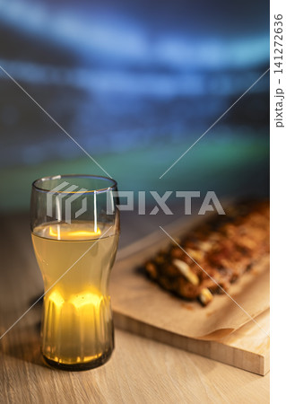 Beverage and grilled ribs on a wooden board arranged for a soccer viewing party, creating a cozy football night atmosphere. 141272636