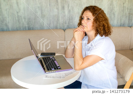 Middle-aged woman working on laptop at home looking thoughtful and concerned about bills 141273179