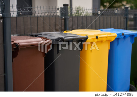 Colorful recycling and waste bins standing in a row beside fence in residential neighbourhood Colorful recycling and waste bins standing in a row beside fence in residential neighbourhood 141274889