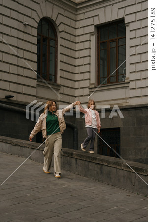Mom and daughter walk along a low wall in a city street on a sunny day in the afternoon Mom and daughter walk along a low wall in a city street on a sunny day in the afternoon 141275189