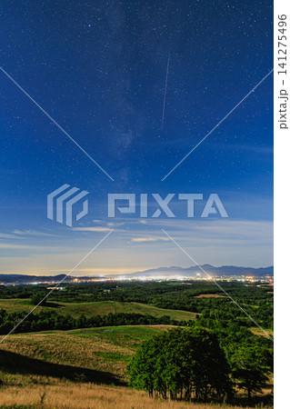 街の夜景を見下ろす広大な丘陵地帯の上に広がる天の川と流れ星の星景風景 街の夜景を見下ろす広大な丘陵地帯の上に広がる天の川と流れ星の星景風景 141275496