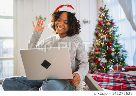 Merry Christmas. African woman in Santa hat having video chat in laptop. Blogger in festive mood recording video in social network. Girl having video call with friends family celebrating Christmas Merry Christmas. African woman in Santa hat having video chat in laptop. Blogger in festive mood recording video in social network. Girl having video call with friends family celebrating Christmas 141275625