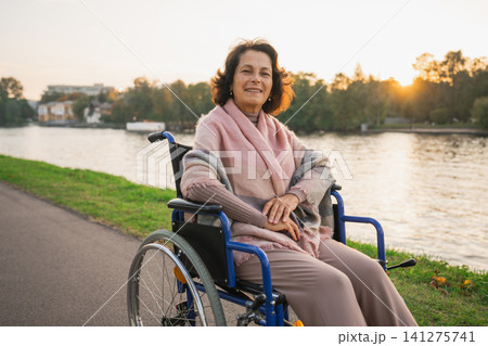 Happy senior woman in wheelchair smiling enjoying freedom walking on road in city park outdoor. Old cheerful positive woman in chair for people with disability. Disability disease rehabilitation Happy senior woman in wheelchair smiling enjoying freedom walking on road in city park outdoor. Old cheerful positive woman in chair for people with disability. Disability disease rehabilitation 141275741