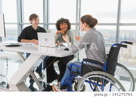 Teamwork brainstorm. Diverse multiracial colleagues woman in chair for people with disability working together. Group of people sitting in creative office discussing business. Young professional team 141275765
