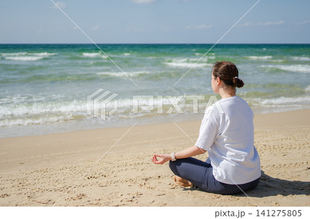 Yoga calm mindfulness meditation. Young healthy woman practicing yoga on beach. Woman sitting in lotus pose meditating relaxing connection with nature by the sea. Girl doing breathing practice Yoga calm mindfulness meditation. Young healthy woman practicing yoga on beach. Woman sitting in lotus pose meditating relaxing connection with nature by the sea. Girl doing breathing practice 141275805