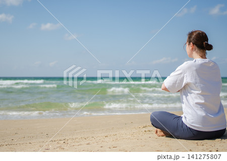 Yoga calm mindfulness meditation. Young healthy woman practicing yoga on beach. Woman sitting in lotus pose meditating relaxing connection with nature by the sea. Girl doing breathing practice Yoga calm mindfulness meditation. Young healthy woman practicing yoga on beach. Woman sitting in lotus pose meditating relaxing connection with nature by the sea. Girl doing breathing practice 141275807