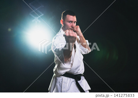 Strong man fighter in white kimono for karate martial arts with black belt preparing for fight training in black background. Man training moves punches kicks practicing shadow fight ready for sparring 141275833