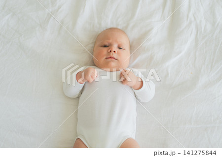 Happy baby. Cute little newborn girl with smiling face lying on bed in bedroom. Infant baby resting playing lying down on white blanket at home. Motherhood happy child childcare concept 141275844
