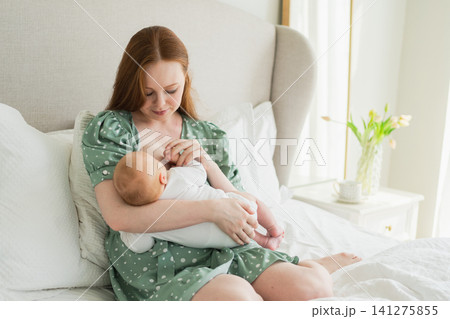 Happy family at home. Mother breastfeeding baby for health nutrition wellness. Mom nursing feeding her newborn child milk at home. Woman infant girl in white bedroom Mother hugs baby with love care 141275855