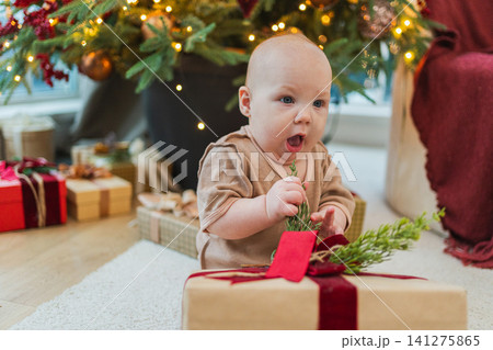 Merry Christmas Xmas. Happy newborn baby child unpacking playing with gift box near Christmas tree at home. Little girl smiling in room with Christmas decorations. Happy baby in first Christmas eve 141275865