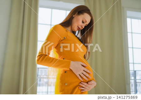 Pregnancy motherhood people expectation future. Pregnant woman with big belly standing near window at home. Girl hugging her tummy enjoying pregnancy. Maternity tenderness parenthood new life concept Pregnancy motherhood people expectation future. Pregnant woman with big belly standing near window at home. Girl hugging her tummy enjoying pregnancy. Maternity tenderness parenthood new life concept 141275869