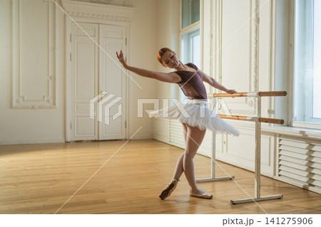 Young ballerina in tutu skirt stretching practicing ballet positions dancing near ballet barre in dance class. Young classical dancer woman in dance studio. Ballet class training Ballet school concept 141275906