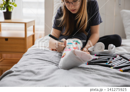 Young teenage girl with broken leg sitting on bed draws with felt tip pens on orthopedic cast. Bored girl with broken leg playing at home during rehabilitation. Accident injury fracture treatment Young teenage girl with broken leg sitting on bed draws with felt tip pens on orthopedic cast. Bored girl with broken leg playing at home during rehabilitation. Accident injury fracture treatment 141275925