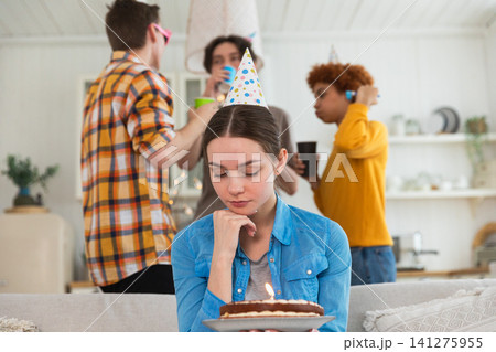 Sad lonely frustrated unhappy young woman looking at birthday cake with candle on birthday party. Girl sad disappointed thinking about life. Birthday party with friends. People celebrating birthday Sad lonely frustrated unhappy young woman looking at birthday cake with candle on birthday party. Girl sad disappointed thinking about life. Birthday party with friends. People celebrating birthday 141275955