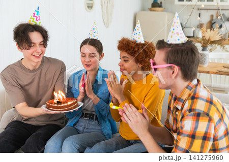 Make a wish. Woman wearing party cap blowing out burning candles on birthday cake. Happy Birthday party. Group of friends wishes girl happy birthday. People celebrating birthday with party at home Make a wish. Woman wearing party cap blowing out burning candles on birthday cake. Happy Birthday party. Group of friends wishes girl happy birthday. People celebrating birthday with party at home 141275960