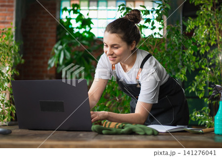 Female florist gardener working in botanical store greenhouse using laptop order plants communicating on Internet with customer. Small business owner working at flower shop. Running of own business Female florist gardener working in botanical store greenhouse using laptop order plants communicating on Internet with customer. Small business owner working at flower shop. Running of own business 141276041