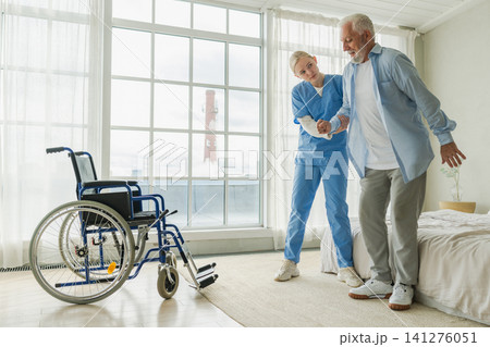 Rehabilitation healthcare. Physiotherapist nurse helps old man patient in wheelchair stand up walking. Man in chair for people with disability doing few steps with nurse helping. Medical caregiver Rehabilitation healthcare. Physiotherapist nurse helps old man patient in wheelchair stand up walking. Man in chair for people with disability doing few steps with nurse helping. Medical caregiver 141276051