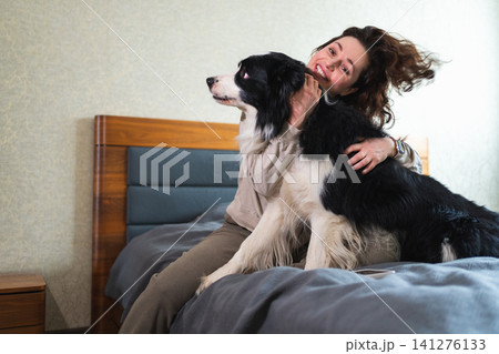 Smiling woman playing with cute puppy dog border collie on bed at home indoor. Owner girl stroking holding dog friend sitting on sofa in living room. Love for pets friendship support team concept Smiling woman playing with cute puppy dog border collie on bed at home indoor. Owner girl stroking holding dog friend sitting on sofa in living room. Love for pets friendship support team concept 141276133