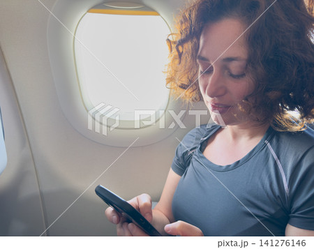 Passenger woman is flying in plane. Girl using phone sitting by airplane window. Traveling female with smartphone inside plane enjoying flight. Woman using wifi connection on board. Traveling girl Passenger woman is flying in plane. Girl using phone sitting by airplane window. Traveling female with smartphone inside plane enjoying flight. Woman using wifi connection on board. Traveling girl 141276146