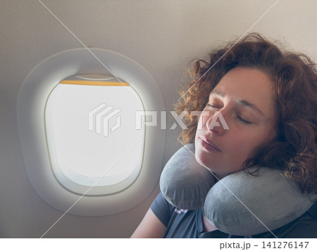Rest on plane. Young woman napping sitting by porthole in business jet using neck pillow for travel. Passenger girl flying in plane. Girl sleeping in airplane going on trip vacation. Traveling girl Rest on plane. Young woman napping sitting by porthole in business jet using neck pillow for travel. Passenger girl flying in plane. Girl sleeping in airplane going on trip vacation. Traveling girl 141276147