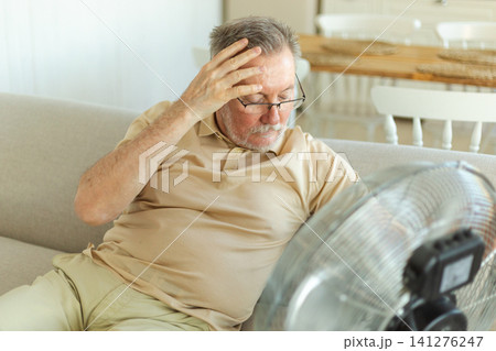 Senior man refreshing himself near fan during summer heat wave hot day. Elderly man suffering from summer heat trying to survive with electric fan. Man enjoying refreshing air blowing from ventilator 141276247