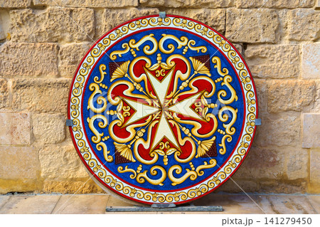 Valletta, Malta - Nov 7 2018, A close up view of a round shield with a colourful image of the Maltese Cross leaning against a brick wall, without people, at daytime, Valletta, Malta Valletta, Malta - Nov 7 2018, A close up view of a round shield with a colourful image of the Maltese Cross leaning against a brick wall, without people, at daytime, Valletta, Malta 141279450
